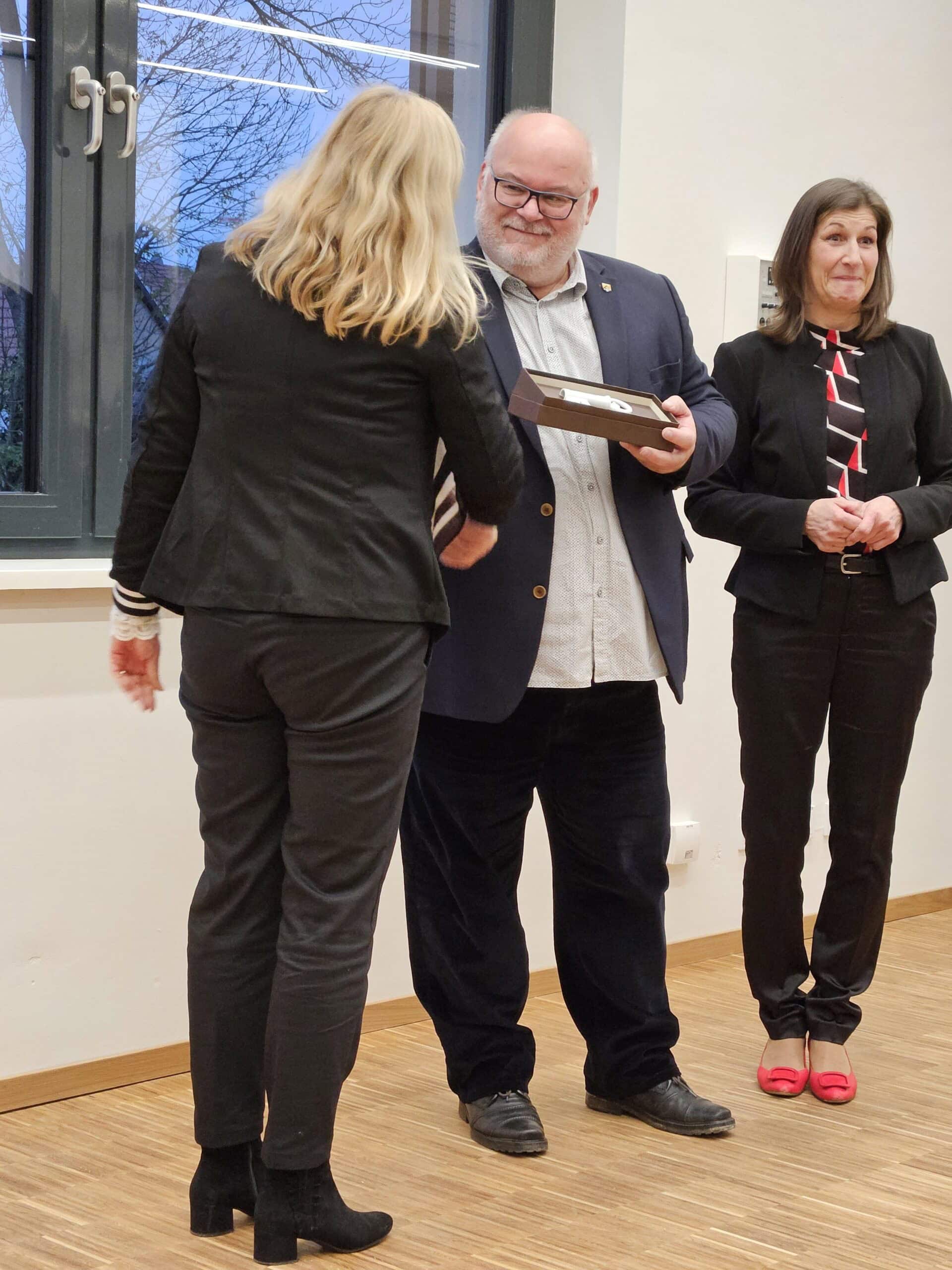 Älterer Herr überreicht einer blonden Frau ein Buch; beide lächeln in einem Raum mit Holzboden.