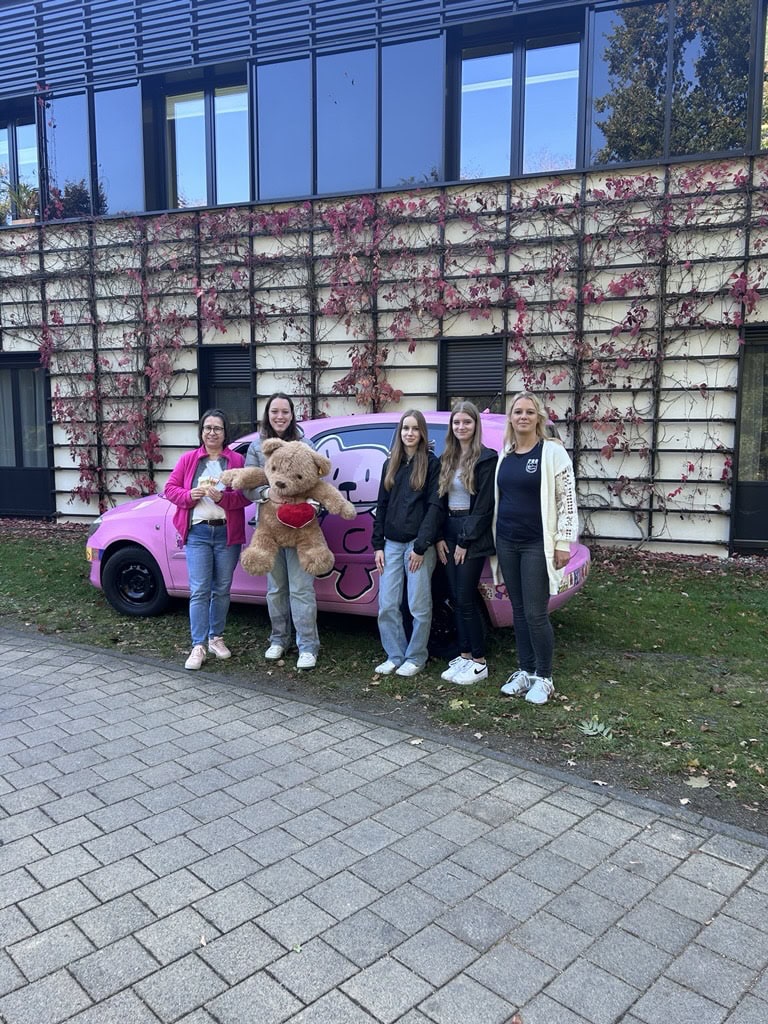 Fünf lächelnde Personen vor rosa Auto mit großem Teddybär, Gebäude im Hintergrund.