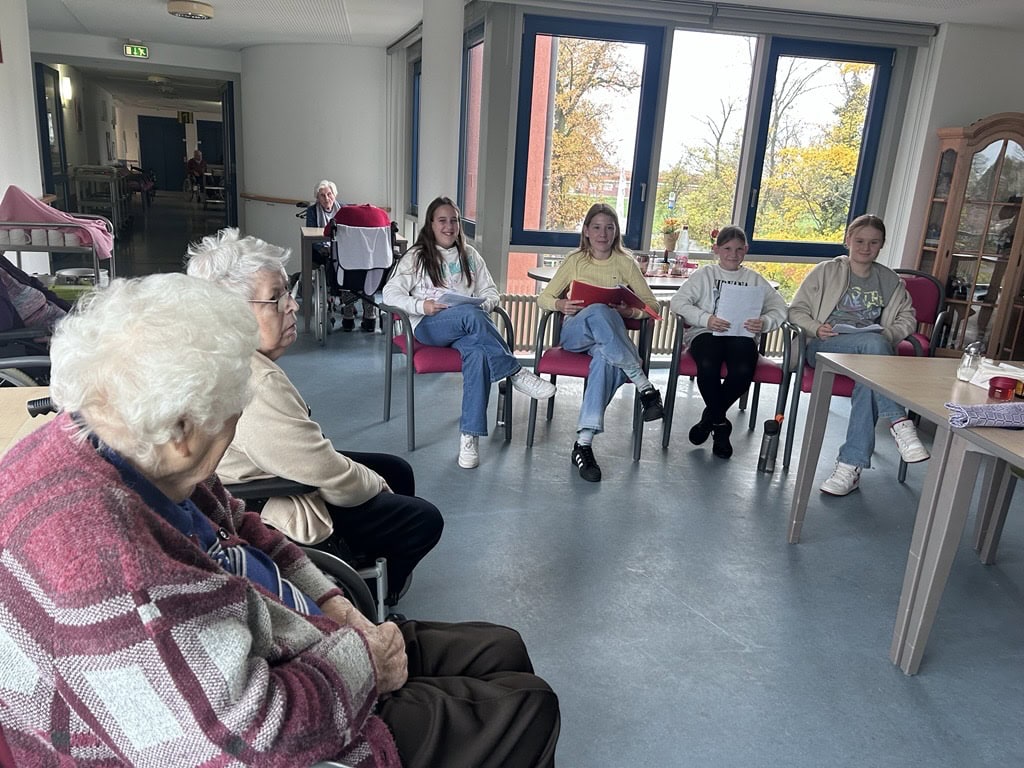 Senioren und Jugendliche in Gesprächsrunde, lächelnd, helles Zimmer mit großen Fenstern.