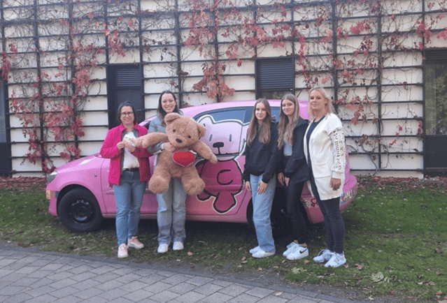 Fünf Frauen lächeln vor einem rosa Auto mit einem großen Teddybär in der Mitte.